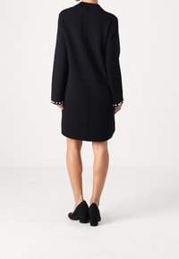 Kvinna iklädd en knälång svart klänning med långa ärmar och pärldetaljer på ärmsluten, tillsammans med svarta klackskor med blockklack, stående mot vit bakgrund.