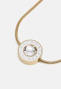 Collana dorata con pendente rotondo, caratterizzata da un cristallo trasparente centrale e dettagli in smalto bianco con la scritta "TOMMY HILFIGER".
