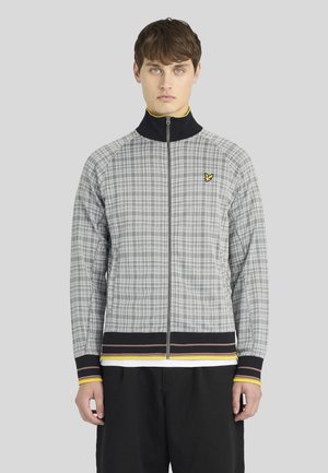 Jeune homme portant une veste zippée à carreaux gris avec un col noir et des poignets rayés, debout devant un fond gris clair uni.