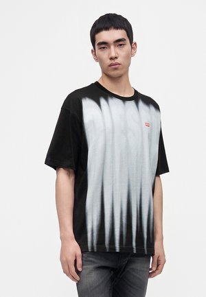 Jeune homme portant un t-shirt dégradé noir et blanc avec un petit logo rouge sur la poitrine, se tenant avec une expression neutre devant un fond blanc.