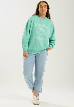 Kvinde iført turkis sweatshirt med palmetrædesign, lyseblå opkørte jeans og hvide sneakers, stående mod en hvid baggrund.