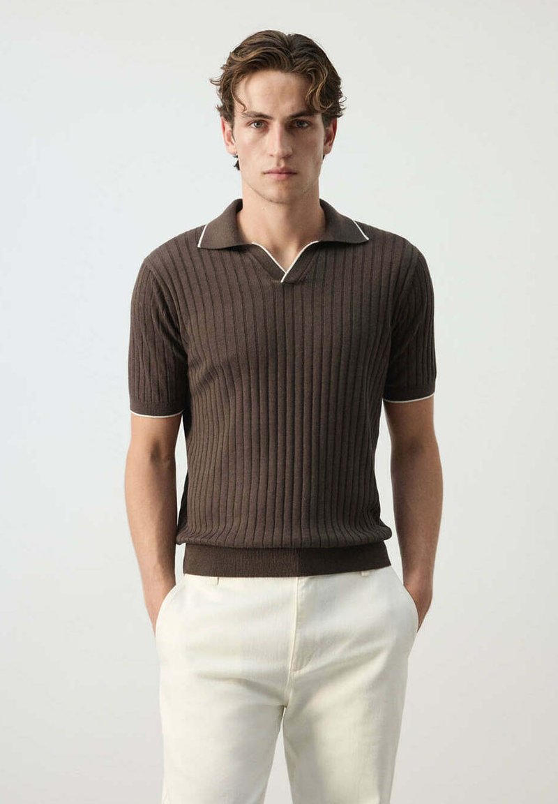 Jeune homme portant un polo marron côtelé à manches courtes et un pantalon blanc, debout, les mains dans les poches, face à un fond uni.