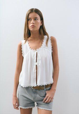 Top sans manches blanc plissé avec dentelle et fermeture à nouer à l'avant. Associé à un short en denim effiloché taille mi-haute. Tissu léger et respirant.