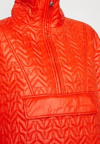 Pikowana, jasnopomarańczowa kurtka z wysokim kołnierzem, przednim zamkiem błyskawicznym i poziomą kieszenią o wyraźnej jodełkowej fakturze.