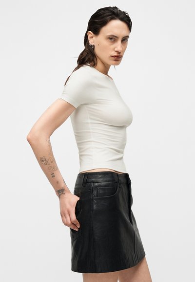 Νεαρό άτομο με σκούρα μαλλιά και τατουάζ, ντυμένο με εφαρμοστό λευκό κοντομάνικο τοπ και μαύρη δερμάτινη φούστα, στέκεται μπροστά σε απλό λευκό φόντο.