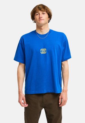 Jonge man met kort bruin haar, gekleed in een blauw T-shirt met een klein geel logo op de borst en donkere broek, staand tegen een witte achtergrond.