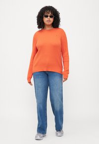 Oranger Strickpullover mit strukturiertem Muster, runder Ausschnitt, gerippte Bündchen, kombiniert mit hellblauen weiten Jeans und silbernen Sneakers.