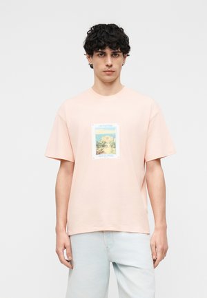 Jeune homme aux cheveux bouclés foncés portant un T-shirt rose clair avec un motif de paysage côtier et un jean bleu clair.