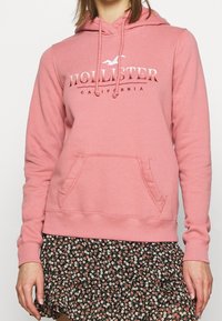 Rosa hoodie i mjukt material, med en frontficka, snoddar och en "Hollister California"-logotyp i en kontrasterande färg.