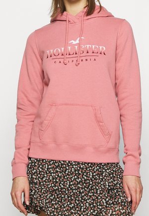 Sudadera rosa hecha de tejido suave, con un bolsillo frontal, cordones y un logo de "Hollister California" en un color contrastante.