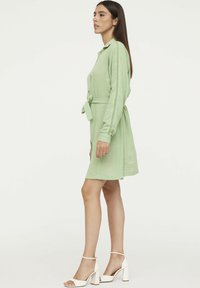 Robe chemise vert clair à manches longues, avec une ceinture à la taille et une poche poitrine boutonnée. Le tissu a un aspect texturé.