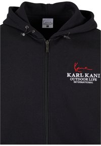 Sudadera con cremallera de color negro hecha de tela suave, con capucha ajustable y logotipo bordado en blanco y rojo en el pecho.
