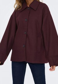 Femme portant un manteau ample boutonné de couleur bordeaux foncé avec de larges manches et un jean bleu foncé, debout devant un fond uni.