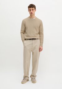 Homme portant un pull beige à côtes, un pantalon beige clair avec ceinture et des chaussures beige à enfiler, debout devant un fond blanc.