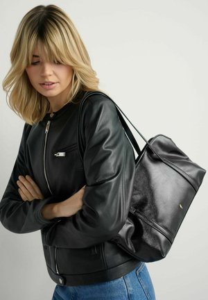 Kobieta w czarnej skórzanej kurtce i dżinsach, niosąca dużą czarną skórzaną torbę na ramię, patrząca w dół z założonymi ramionami.