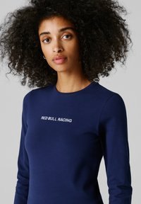 Ženska s kodrastimi lasmi, oblečena v temno modro majico z dolgimi rokavi, na prsih ima napis "RED BULL RACING", na preprostem sivem ozadju.
