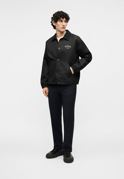 Νέος άνδρας με μαύρο σακάκι με κουμπιά και λογότυπο, μαύρο παντελόνι και μαύρα αθλητικά παπούτσια, στέκεται με το ένα χέρι στην τσέπη, κοιτώντας προς τα αριστερά.