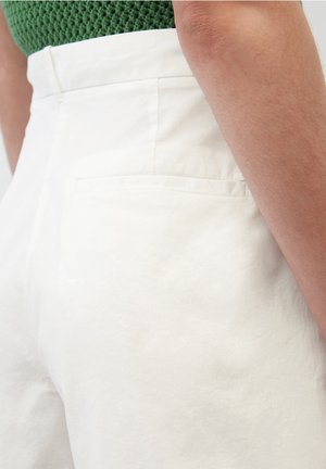 Pantalons blancs taille haute en tissu léger, dotés d'une poche arrière discrète et d'une texture lisse, présentés sur un fond en tricot vert.