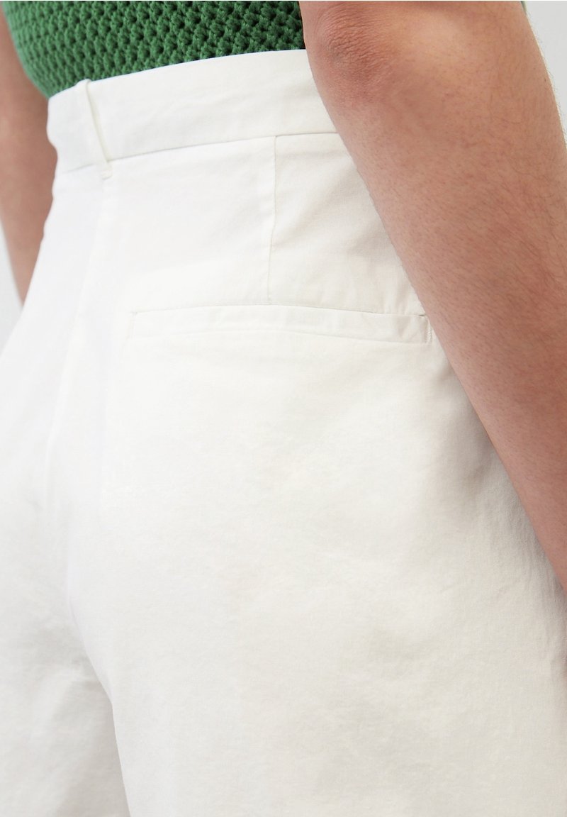 White high-waisted pants made of lightweight fabric, featuring a discreet back pocket and smooth texture, shown against a green knit background.