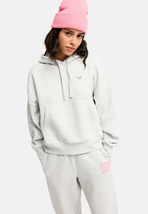 Jeune femme portant un sweat à capuche gris clair et un pantalon de survêtement avec un texte rose, ainsi qu'un bonnet tricoté rose, debout les mains dans les poches.
