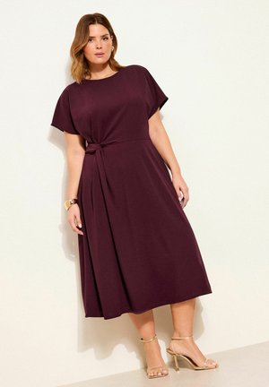 Frau trägt ein burgunderrotes Midikleid mit kurzen Ärmeln und seitlicher Schleife, steht vor einer hellen Wand und trägt goldene Riemchenabsätze sowie ein Armband.