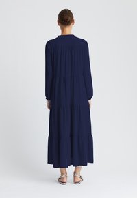 Marineblaues Maxikleid mit langen Ärmeln, Stufendesign und gerafften Details. Glatter Stoff und lockere Passform, kombiniert mit Riemchensandalen.