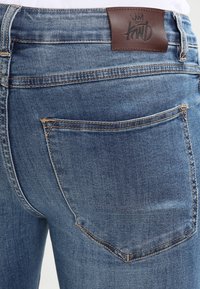 Närbild av blå denimjeans bakfickan med en brun läderlogotyp på midjan och synliga sömdetaljer.