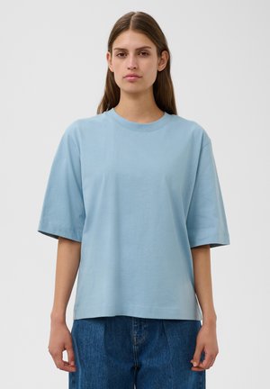 Jeune femme aux longs cheveux bruns portant une chemise bleu clair oversize à manches courtes et un jean bleu, debout devant un fond blanc uni.