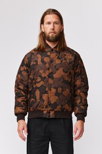 Brun camo puffer-jakke med en quiltet tekstur, der har en trykknap-lukning fortil og ribbede manchetter. Designet inkluderer jordfarver i brun og sort.