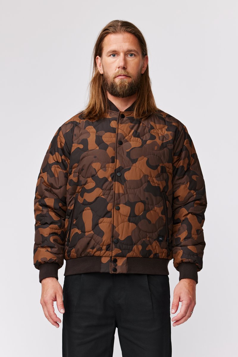 Brun camo puffer-jakke med en quiltet tekstur, der har en trykknap-lukning fortil og ribbede manchetter. Designet inkluderer jordfarver i brun og sort.