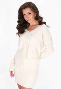 Elfenbeinfarbene Strickkleid mit V-Ausschnitt, langen Ärmeln und dekorativen Perlen. Weicher Stoff, figurnah in der Taille und bis zur Mitte des Oberschenkels.