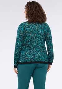 Pulover cu model turcoaz, având designuri zigzag în alb și negru, cu o bandă neagră la tiv și la manșete; purtat cu pantaloni verzi.