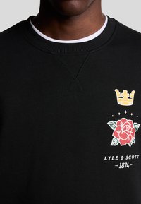 Schwarzer Pullover mit Rundhalsausschnitt, verziert mit gelber Krone und roserosenbestickung sowie dem Text "LYLE & SCOTT - 1874" am unteren Ende.