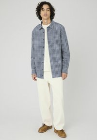 Chemise à carreaux bleu marine et blanche, en coton, coupe décontractée, portée sur un haut crème, assortie à un pantalon crème et des chaussures marron.