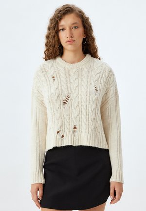 Pull en tricot épais de couleur blanc cassé avec des poignets et un ourlet côtelés. Présente des découpes dans un design court, associé à une jupe noire.