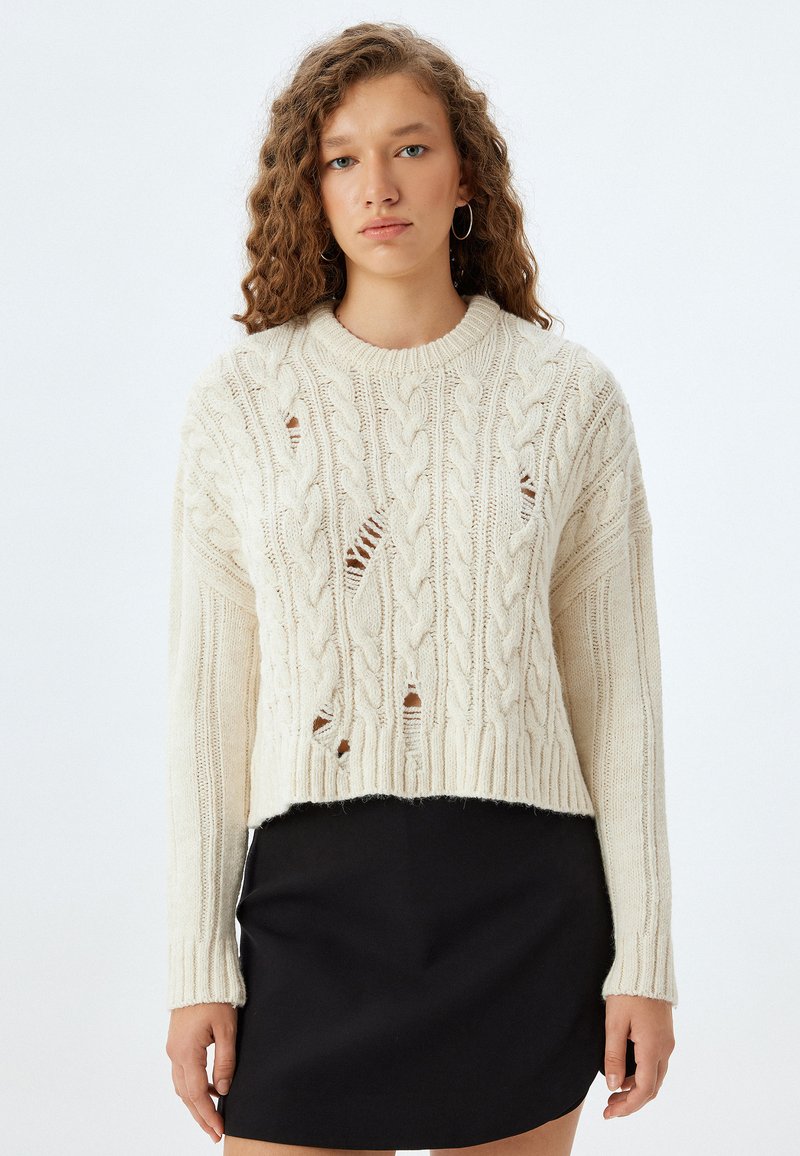 Dikke kabelgebreide trui in off-white met ribgebreide manchetten en onderrand. Heeft uitsparingen in een cropped ontwerp, gecombineerd met een zwarte rok.
