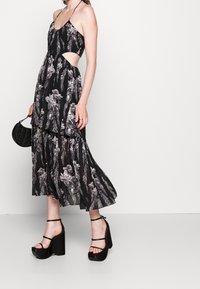 Nainen mustassa kukallisessa halterneck-mekossa, jossa on leikkuusivut, mustissa korokepohjaisissa sandaaleissa, pitämässä pientä mustaa päällikkölaukku.