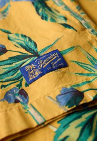 Tejido amarillo con un estampado botánico que presenta hojas verdes y flores azules. Un etiquetado azul está adjunto con texto dorado y un gráfico de una palmera.