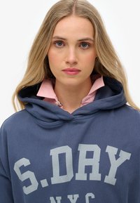 Sudadera navy con letras grises "S.DRY NYC," con capucha ajustable y una camisa de color rosa pálido con cuello debajo. Textura suave.