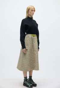 Pulôver preto, saia midi acolchoada caqui com bolsos laterais, e cinto amarelo. Modelo a usar sapatos cinzentos e meias até ao tornozelo.