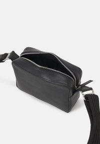 Sac en cuir noir porté en travers avec une large sangle réglable, fermeture éclair partiellement ouverte, montrant un intérieur vide sur fond blanc.