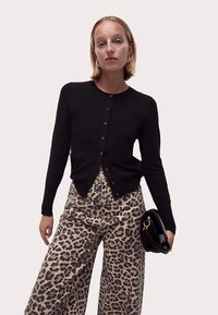 Svart stickad kofta med knappar, kombinerad med vida byxor i leopardmönster. Modellen håller en svart handväska som visar en mjuk textur.