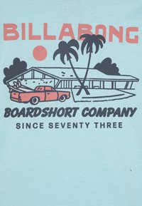 T-shirt blu chiaro con una grafica di un camion rosso d'epoca, una casa moderna, palme, e il testo "BILLABONG" e "BOARDSHORT COMPANY."