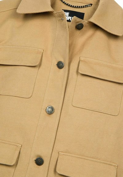 Chemise utilitaire beige avec un devant boutonné, deux poches à rabat et des boutons-pression noirs contrastants. Le tissu a un aspect texturé.