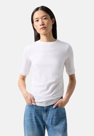 Young woman with long dark hair stands with hands in pockets wearing a plain white short-sleeve top and blue jeans on a white background.