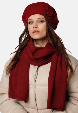 Berretto di maglia rosso e sciarpa coordinata abbinati a un giubbotto trapuntato color crema, caratterizzato da una texture morbida e dettagli floreali sul cappello.