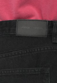 Fekete farmernadrág sima textúrával, melyen egy téglalap alakú fekete bőr címke található, rajta a "SAMSEØ SAMSEØ" dombornyomott felirattal és varrási részletekkel.