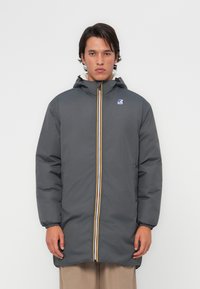 Veste grise rembourrée avec capuche, dotée d'une fermeture éclair sur le devant accentuée par des rayures jaunes et blanches ainsi qu'un logo sur le haut à gauche.