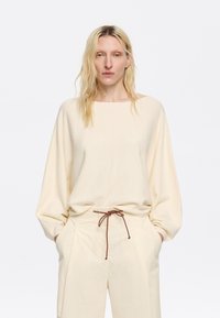 Maglioncino in pile crema con scollo ampio e maniche lunghe a palloncino, abbinato a pantaloni di colore chiaro con un laccio marrone.