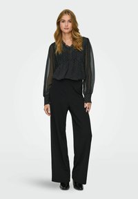 Femme debout portant un pantalon noir à jambes larges et un chemisier noir transparent à manches longues avec des pois blancs, devant un fond uni.
